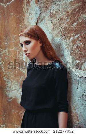 Portrait of young beautiful red-haired girl in trendy black dress posing over graphic old wall. Perfect skin and strong smoky eyes make-up. Glazed hair. Hipster style. Indoor shot