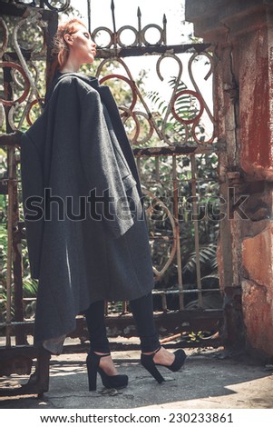 Retro vogue fashion concept. Portrait of young beautiful red-haired girl in trendy grey coat posing near old garden and ruin gate. Perfect skin and strong smoky eyes make-up. High fashion style.