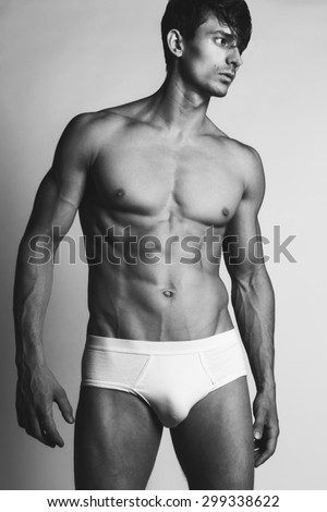 Male swimwear & underwear concept. Handsome muscular male model in trendy white underwear posing over grey background. Black & white photo. Studio shot
