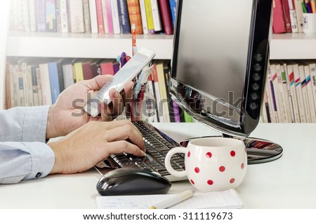 Side view shot of a man\'s hands using smart phone and computer at  office desk.