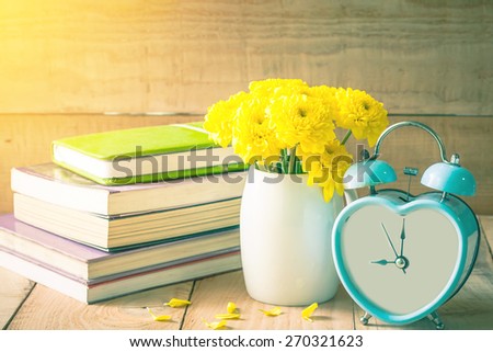 soft focus of old retro clock on wooden floor with yellow flower in white pot and old books background, vintage color tone.