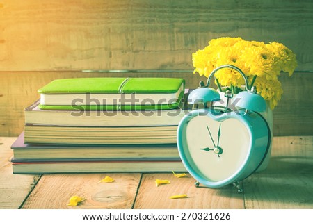 soft focus of old retro clock on wooden floor with yellow flower in white pot and old books background, vintage color tone.
