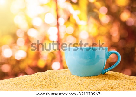 blue cup of coffee or tea on sand over autumn forest with sun lighting background.
