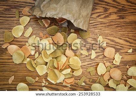 Closeup view from above of colorful veggie potato, tomato, spinach chips falling out of brown paper bag on a wooden table, natural clean healthy vegan cooking eating concept