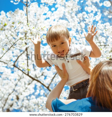 Mother holding her son boy toddler high in her arms, blooming white tree flowers and blue sky on the background, toddler smiling laughing showing his tongue being funny