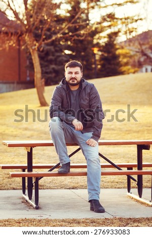 Portrait of a young sexy bearded white Caucasian man in jacket, jeans sitting on the wooden bench table outside in park at sunset, looking at something interesting in front of him, thinking