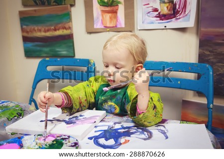 Cute adorable little baby boy girl toddler sitting in art studio indoors and drawing painting with brushes, pencils, pastel. Early child development concept