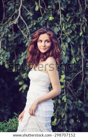 Portrait of beautiful innocent Caucasian adult girl woman with long dark red brown hair in white summer open dress in park outside among green trees plants on sunset