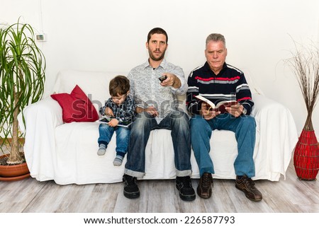 baby, father and grandfather watching tv, while baby is playing with tablet and grandfather is reading a book