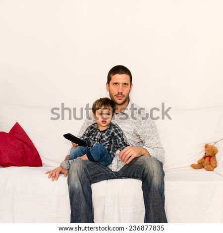 Bored father watching tv with his son sitting on his knees