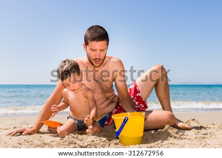 close-up of cute and smiling caucasian father and son playing with sand, pail and shovel on the beach with sea background during summer
