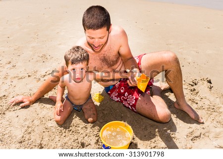 close-up of cute and smiling caucasian father and son playing with sand, pail and shovel on the beach during summer