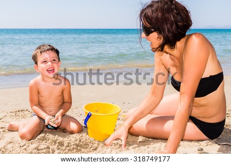 close-up of cute and smiling caucasian mother and son playing with pail and shovel on the beach with sea background during summer