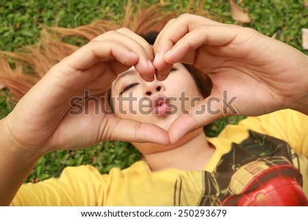 woman lies down on green grass showing hands in heart-shaped with kiss mouth behind