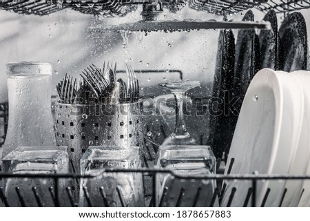 Transparent and black and white dishes as well as cutlery and glasses are washed in the dishwasher, inside view, drops and splashes of water.