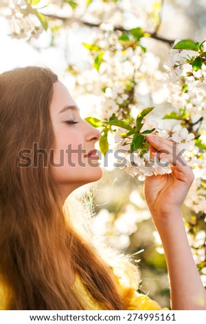 Young smiling girl holds the branch of cherry tree with white flowers and smells the tender smell of flowers, on a warm and sun spring background.