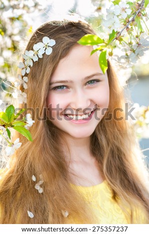 Young smiling girl with white cherry flowers in her long hairs looks ahead on a background with spring trees and white flowers. Natural beauty and color.