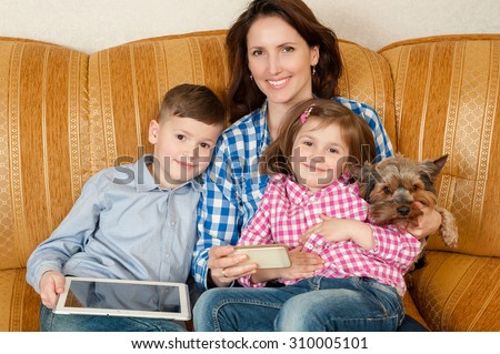 Happy mother with the son and the daughter on a sofa. The son holds the digital tablet, mother embraces the daughter and holds the mobile phone and a dog.