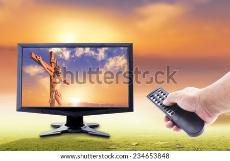 Human hands holding remote and monitor display Jesus christ with the cross on sunset over the actual location.