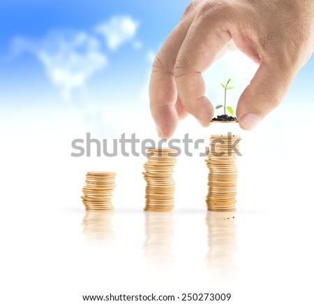 Human hand adding a golden coin with young plant in the final row over blurred world map of clouds background. Money coin concept.