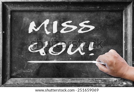 Human hand holding chalk and writing text for MISS YOU! on blackboard.
