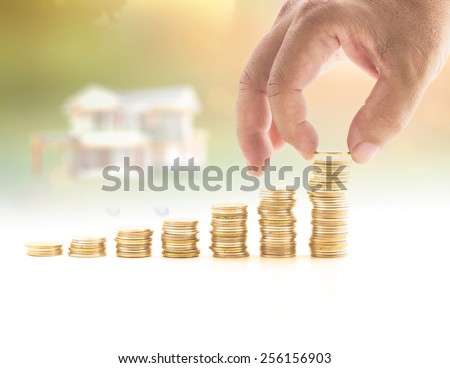 Human hand adding a golden coin in the final row of golden coins over blurred house on sunset background. Concept for money coin, insurance, buying, renting, service.