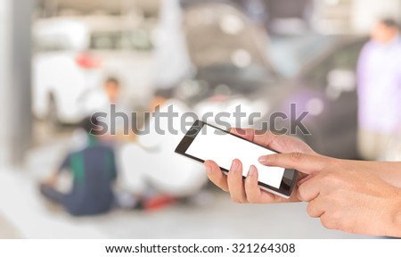 male hand is holding a modern touch screen phone and blur image of worker fixing car in the garage for background usage.