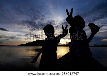 Silhouetted kids ( excited,joyous sister holding an energy drink and uninterested, not so happy younger brother) showing peace sign with their hand/finger during sunset