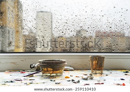On the windowsill is palette with oil paints and dirty glasses. Rain at the behind the window. water drops against gray spring sky with buildings. focus sat at the bottom.