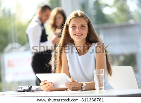 Smiling attractive woman student sitting at the table with a tab and  other students standing in background