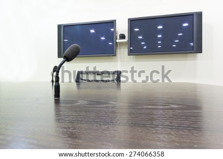 Black microphone on a wooden brown table in a city conference room, with two TV screens with light reflections in the background