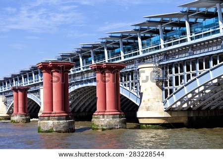 View from south bank of west side of arch wrought iron London Blackfriars Railway Bridge with platforms crossing River Thames in London along old red pillars, solar panels powered