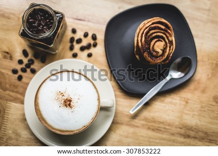 Cup of coffee and bun with cinnamon lying on the wooden table and coffee bean in cafe flower nature background.