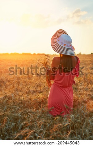 Beautiful young woman with brown hear wearing rose dress and hat  looking to the sun on perfect wheat field on sunset