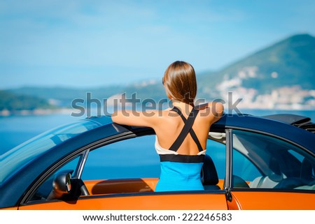 Beautiful young woman with long hair standing back with orange cabriolet at the Mediterranean sea coast