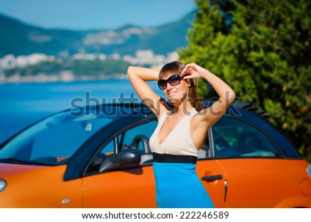 Beautiful young woman with long hair standing near orange cabriolet at the Mediterranean sea coast