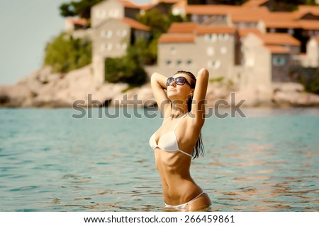 Sexy beautiful woman in white bikini with creative white sunglasses on Mediterranean sea coast. Sveti Stefan, Montenegro, South Europe. Retro toned image