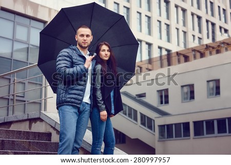 Beautiful young couple in love on a date outdoors on modern urban background. Bearded handsome man and brunette pretty woman in casual dress with umbrella. Rainy weather.