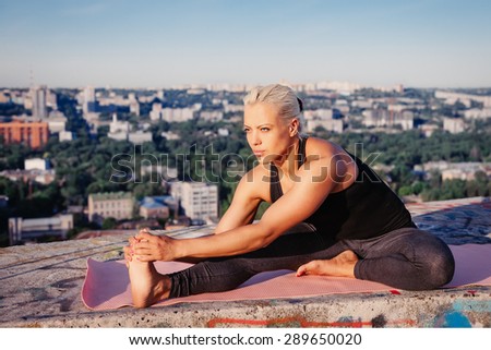 Portrait of blonde strong woman in sportwear doing workout stretching on the roof of skyscraper above the big city at early morning. Female sporty muscular body. Dawn and sunrise bright sunny light.