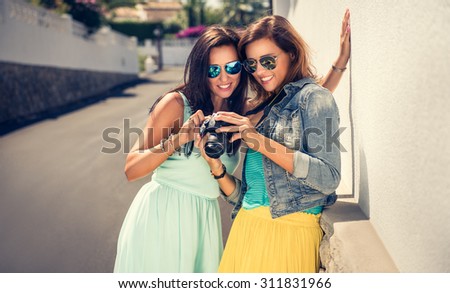 Outdoor lifestyle fashion portraits of two sisters in modern, colorful clothes, with digital camera. Two best friends hugs and posing, having fun on a sunny day. Women looking at the camera.