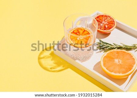Half of grapefruit and bloody orange, rosemary on a white wooden tray and glass with water on bright yellow background. Summer refreshment concept. Sunlit flat lay. Minimal style. Top view.