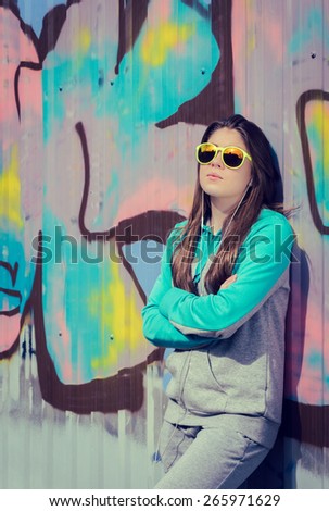 Stylish teenage girl in colorful sunglasses posing near graffiti wall