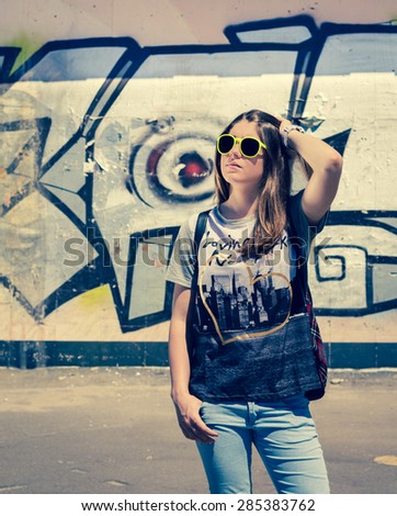 Stylish teenage girl in sunglasses posing near graffiti wall in a city in a summer day. Toned effect