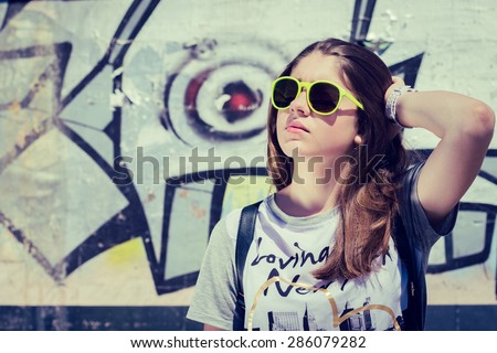 Portrait of a stylish teenage girl in sunglasses posing near graffiti wall in a city in a summer day. Toned effect