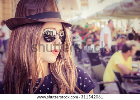Portrait in profile of a stylish girl in a hat and sunglasses with street restaurant in the background