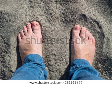 Bare feet on dark sand. Top view. Ugly feet with orange-pink nail polish seen from the top. Part of jeans visible. Person is standing on the beach. Feet look tired, maybe the shoes where too small.