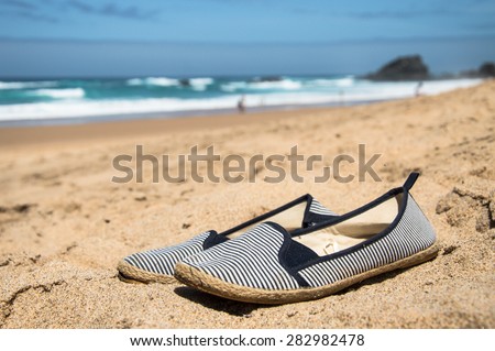 Summer shoes on the beach. Summery, navy blue and white stripes sneakers on the golden sand on blurred background (wavy sea, sky, some rocks). Nautical, sailor style sneakers. Summer holidays feeling.