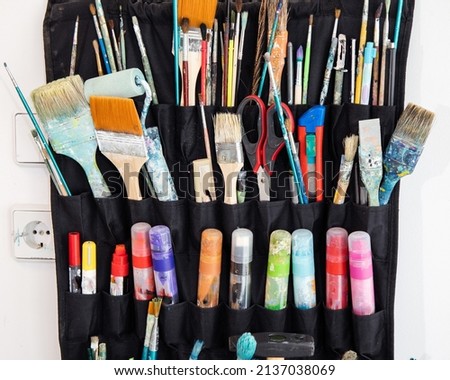 Closeup of multiple colorful dirty paintbrushes, pens, pencils, markers and other artistic tools organized in rows inside black holder hanging on the white wall