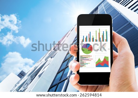 hand holding mobile phone with analyzing graph against office buildings with sun