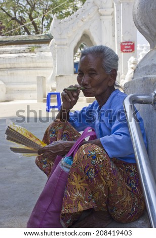 Mingun, Myanmar - February 28, 2014: Old Asiatic woman smoking a cigar outside a temple. The old woman sells fans and cigars to the tourist outside the religious place.